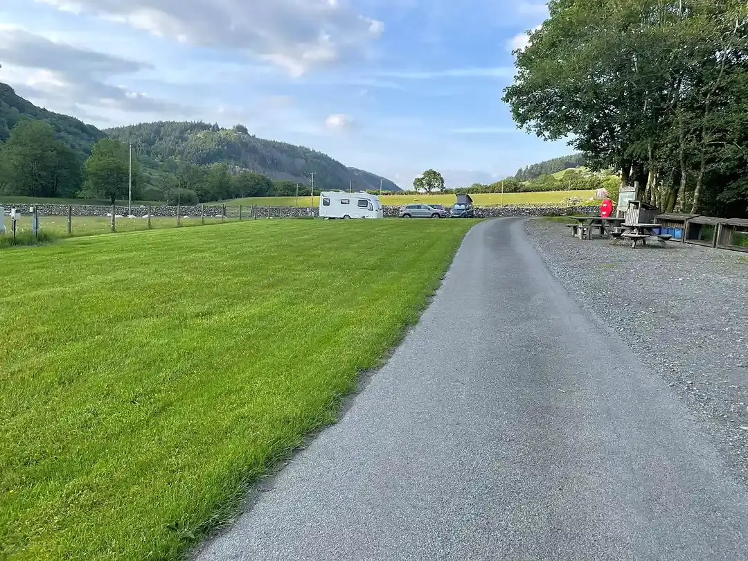The smooth tarmac lane through Tŷ Cerrig with a caravan pitched on the grass ahead and hills in the distance